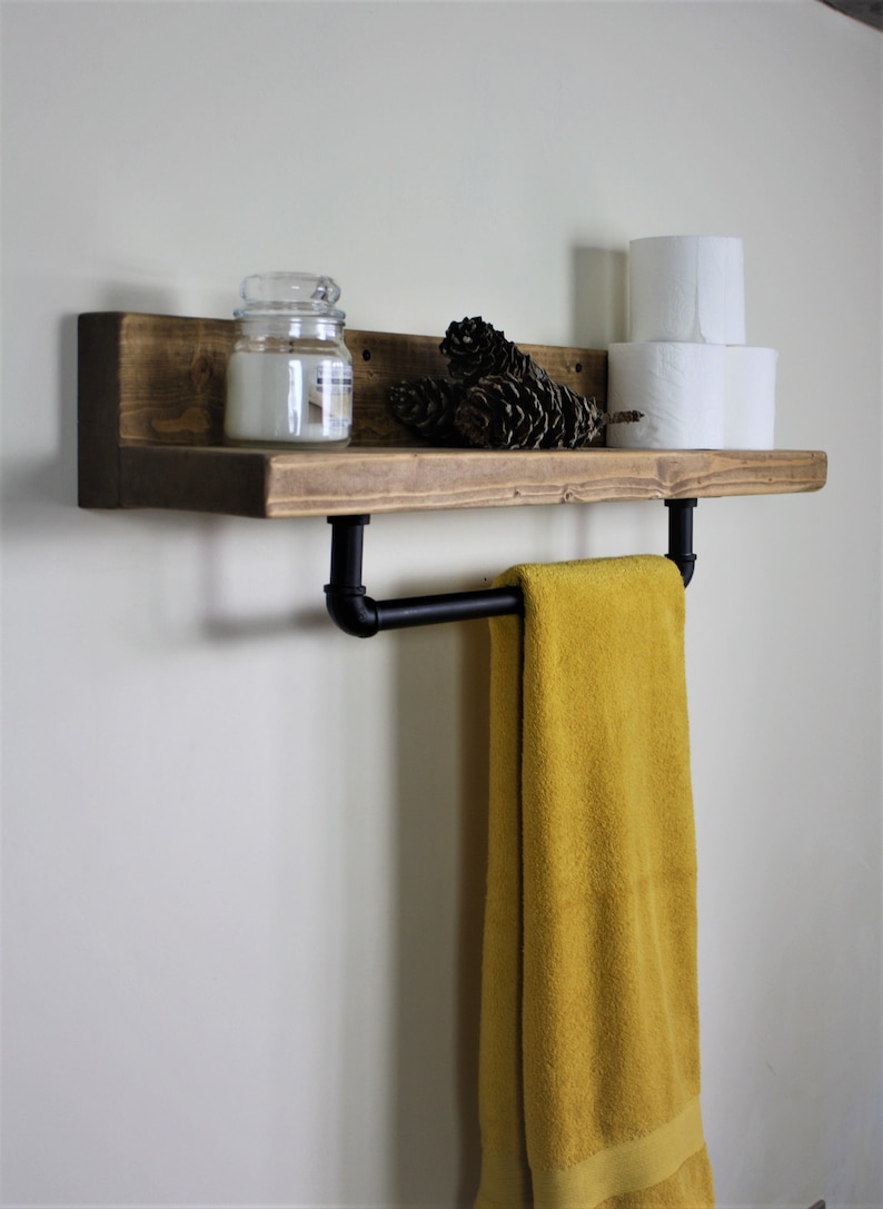 Pu&ograve; includere: Uno scaffale in legno rustico con un portasciugamani in tubo di metallo nero. Lo scaffale &egrave; decorato con un barattolo di vetro, pigne e due rotoli di carta igienica.