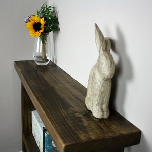 May include: A dark brown wooden shelf with a white ceramic rabbit figurine and a glass vase with yellow sunflowers on top. The shelf has two lower shelves with books on them.