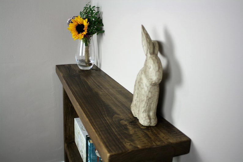 May include: A dark brown wooden shelf with a white ceramic rabbit figurine and a glass vase with yellow sunflowers on top. The shelf has two lower shelves with books on them.