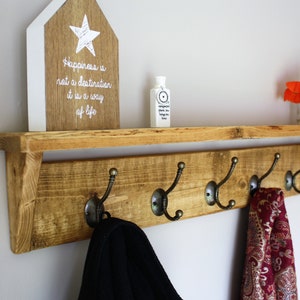 Peut inclure: &Eacute;tag&egrave;re en bois rustique avec cinq crochets en m&eacute;tal, une petite pancarte en bois avec le texte "Le bonheur n'est pas une destination, c'est un mode de vie", et une petite bouteille sur l'&eacute;tag&egrave;re. Il y a des fleurs rouges dans un vase en verre sur l'&eacute;tag&egrave;re.