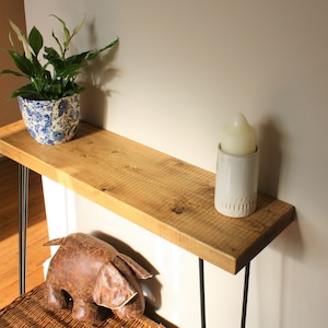 May include: A wooden console table with black hairpin legs. The table is decorated with a potted plant, a candle in a white holder, and a brown stuffed elephant.