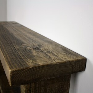 May include: Close-up of the top of a rustic wooden bench with a dark brown stain. The bench has a thick, rounded top and is made of rough-hewn wood.