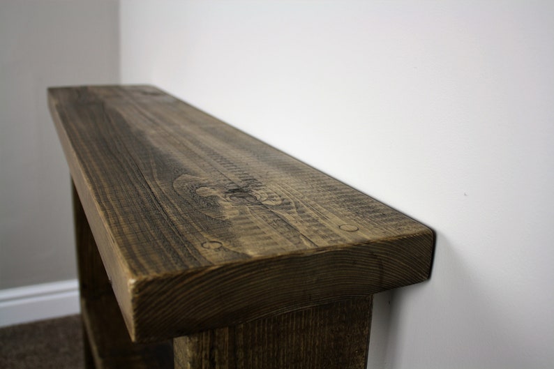 May include: Close-up of the top of a rustic wooden bench with a dark brown stain. The bench has a thick, rounded top and is made of rough-hewn wood.