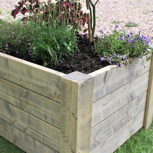 May include: A wooden planter box with a weathered grey finish. The planter is filled with soil and various plants, including purple flowers and green foliage.