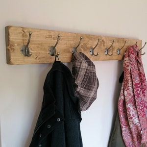 May include: A rustic wooden coat rack with six antique-style hooks. A black jacket, a plaid hat, and a patterned scarf are hanging on the hooks.