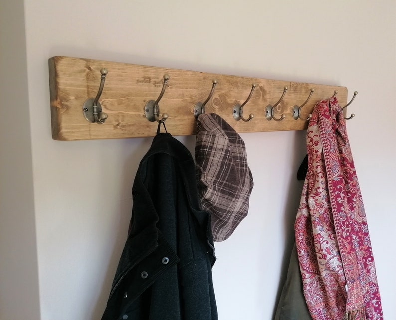 May include: A rustic wooden coat rack with six antique-style hooks. A black jacket, a plaid hat, and a patterned scarf are hanging on the hooks.