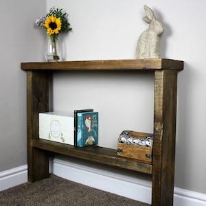 May include: A rustic wooden console table with a single shelf. The table is stained a dark brown color and has a distressed finish. The table is decorated with a vase of sunflowers, a ceramic rabbit figurine, and a few books.