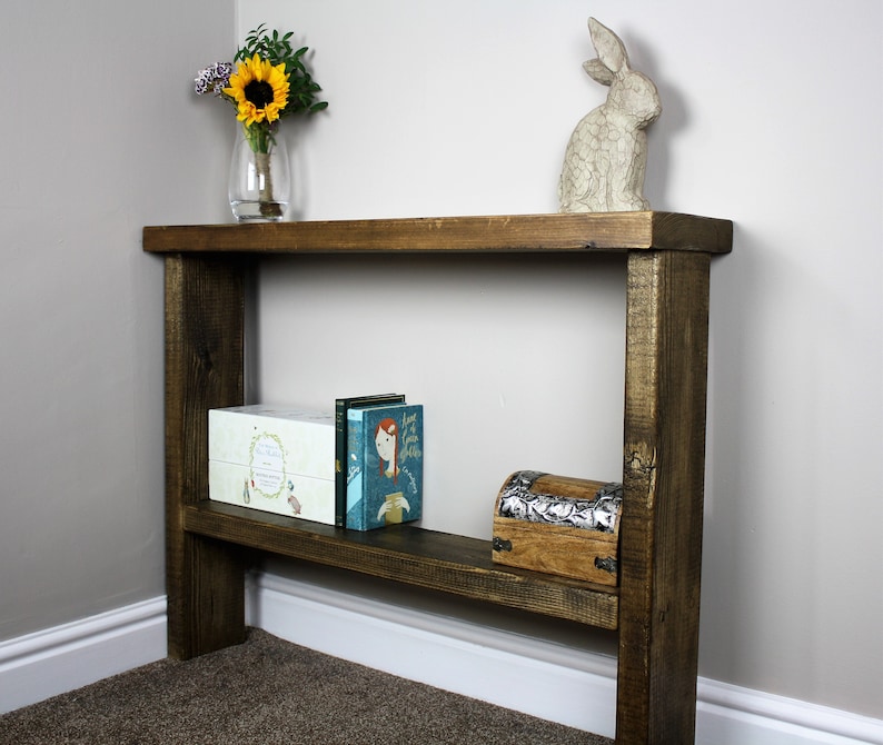 May include: A rustic wooden console table with a single shelf. The table is stained a dark brown color and has a distressed finish. The table is decorated with a vase of sunflowers, a ceramic rabbit figurine, and a few books.