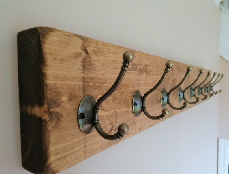 May include: A rustic wooden coat rack with eight antique brass hooks. The rack is stained a dark brown color and has a natural wood grain.