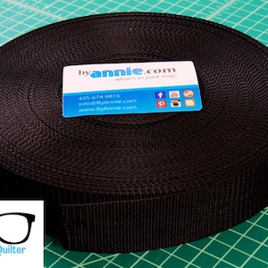 May include: A roll of black nylon webbing, approximately 1 inch wide. The webbing is rolled up and a business card is on top of the roll. The business card has the text "byannie.com" and a phone number, email address, and website address.