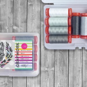 May include: Two clear plastic sewing thread storage boxes. One box features a wolf head design and the text "GRAYWORK". The other box contains spools of thread in various shades of gray, white, and navy blue.