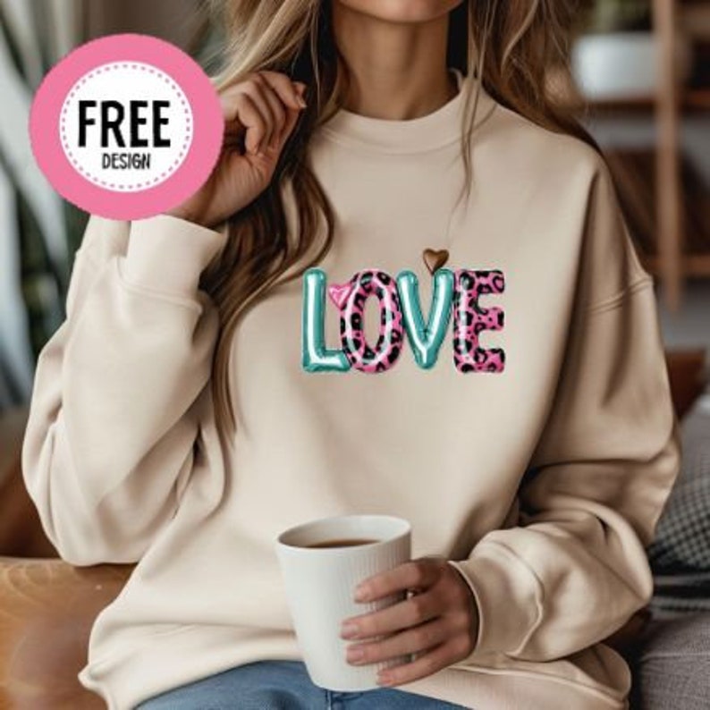 Może przedstawiać: Kremowa bluza z nadrukiem słowa "LOVE" z przodu. Litery są w jasnoniebieskim kolorze z r&oacute;żowym nadrukiem w panterkę. Małe złote serce znajduje się nad słowem.