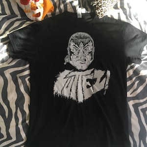 Puede incluir: Camiseta negra con un estampado gráfico blanco de un luchador enmascarado con la palabra "MIEZ" debajo.