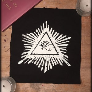 May include: A black fabric patch with a white all-seeing eye design inside a triangle with a radiating sunburst pattern.