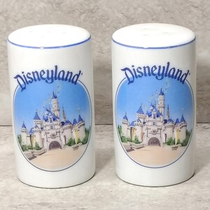 May include: A pair of white ceramic salt and pepper shakers with a blue rim. Each shaker has a Disneyland logo and a colourful image of a castle with blue turrets against a blue sky.