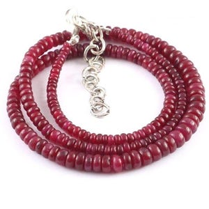 May include: A layered necklace featuring small, round, deep red beads. The beads are strung together, forming multiple strands. A silver-colored chain and clasp are visible at the top, allowing for adjustable length. The necklace is displayed against a white background.