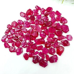 Puede incluir: Una colección de piedras preciosas de rubí en bruto, sin cortar, de varios tamaños y formas. Las piedras preciosas son de un color rojo intenso y rico y parecen translúcidas. Los rubíes están dispersos sobre una superficie blanca, con hierba decorativa verde en el fondo.