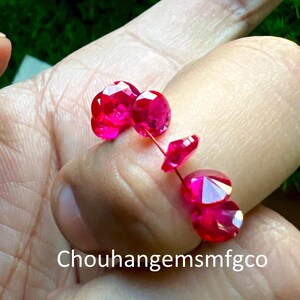 May include: Close-up of several faceted, bright red gemstones. The gems are held in a hand, showcasing their brilliant cut and color. The background is a blurred green, providing contrast to the gems. Text: Chouhangemsmfgco.