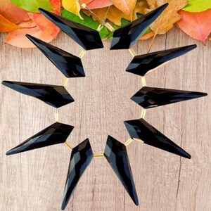 May include: A striking black necklace featuring geometric, triangular pendants. The pendants are connected by a gold-colored chain, creating a bold and modern design. The necklace is displayed against a wooden background with colorful leaves.