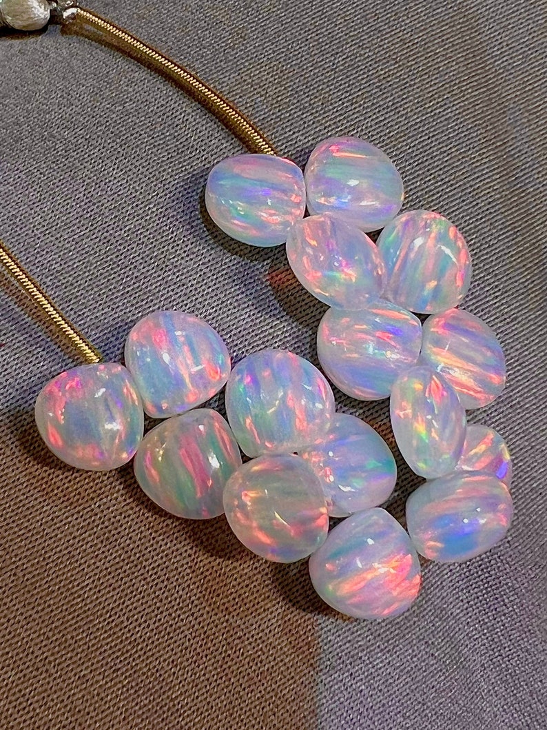 May include: A collection of polished, iridescent opal gemstones. The opals display a play of color with flashes of pink, blue, and green. They are smooth and rounded, with a pearly white base color. A gold chain is visible in the upper left corner.