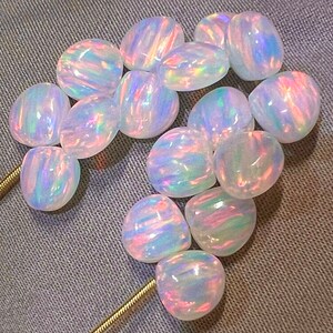May include: A collection of polished, oval-shaped opals. The opals have a milky white base color with iridescent flashes of pink, blue, and green. The gemstones are arranged on a gray fabric surface, with a gold chain visible.