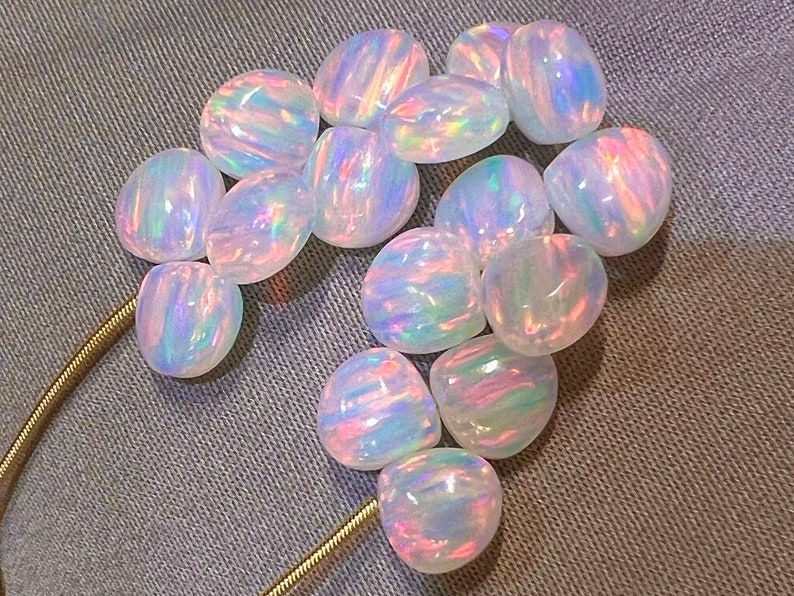May include: A collection of polished, oval-shaped opals. The opals have a milky white base color with iridescent flashes of pink, blue, and green. The gemstones are arranged on a gray fabric surface, with a gold chain visible.