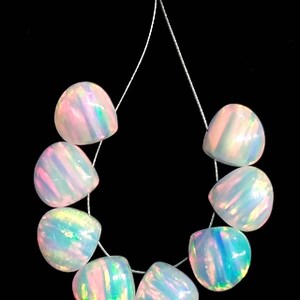 May include: A strand of eight iridescent opal beads. Each bead is a rounded, irregular shape with a pearlescent sheen, displaying flashes of pink, blue, and green. The beads are strung on a thin, clear cord against a black background.