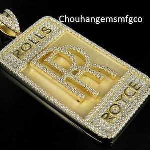 May include: A gold-colored pendant with the Rolls Royce logo, encrusted with small, clear gemstones. The rectangular pendant has the word "ROLLS" at the top and "ROYCE" at the bottom. The pendant is on a black background.