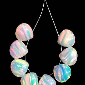 May include: A strand of eight iridescent opal beads. Each bead is a rounded, irregular shape with a pearlescent sheen, displaying flashes of pink, blue, and green. The beads are strung on a thin, clear cord against a black background.