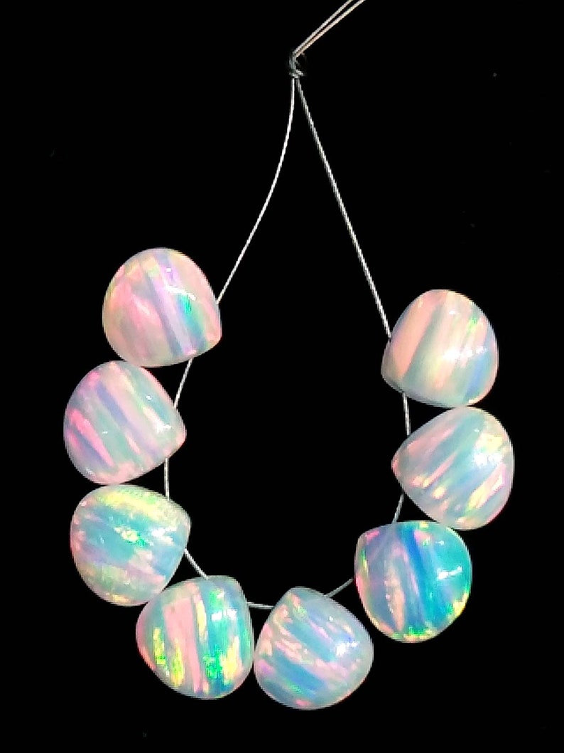 May include: A strand of eight iridescent opal beads. Each bead is a rounded, irregular shape with a pearlescent sheen, displaying flashes of pink, blue, and green. The beads are strung on a thin, clear cord against a black background.