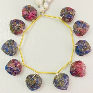 May include: A strand of eight heart-shaped carved gemstones featuring a gradient of blue, purple, and pink hues with gold-colored metallic accents. The stones are strung on a thin gold-colored wire and finished with a white tassel at the top.