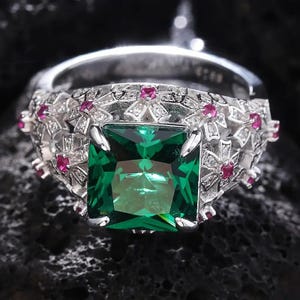 Peut inclure: Une bague en argent ornée d'une grande pierre précieuse vert émeraude de taille carrée. La bague est ornée de petites pierres précieuses rondes rouge rubis et de détails en filigrane argenté. La bague est posée sur un fond sombre et texturé.