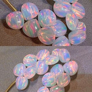 May include: A collection of iridescent opal gemstones. The opals are oval-shaped, displaying a play of color with hues of pink, blue, and green. They are arranged on a textured gray surface, with a gold chain visible.