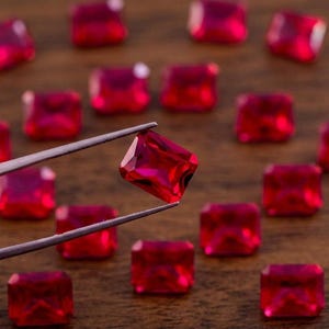 May include: Close-up of several faceted, rectangular, bright red gemstones. One gemstone is held by silver tweezers, showcasing its clarity and cut. The gems are scattered on a warm-toned wooden surface, creating a contrast of color and texture.