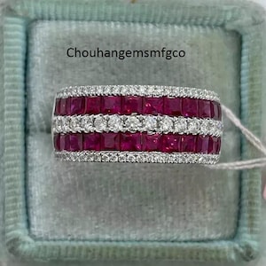 Può includere: Un anello largo, color argento, con tre file di pietre preziose rosse scure a taglio quadrato, intervallate da due file di pietre preziose chiare, rotonde e più piccole. L'anello è posto su uno sfondo azzurro chiaro. Il testo "Chouhanggemsmfgco" è visibile.