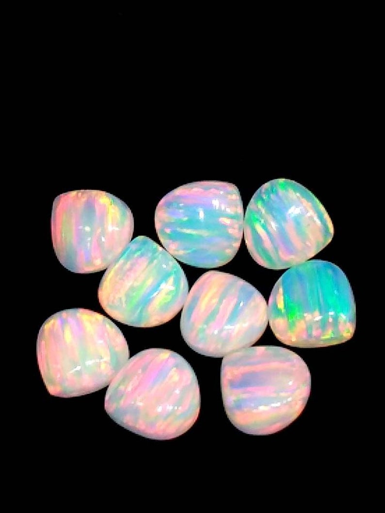 May include: Nine polished opal gemstones with a pear-like shape. The opals display a range of colors, including pink, blue, green, and yellow, with a shimmering, iridescent effect. The gemstones are set against a black background.