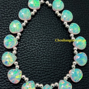 May include: Strand of heart-shaped opals with iridescent colors of green, blue, and pink. The opals are strung together with small, silver-colored beads. The background is a dark, textured surface.