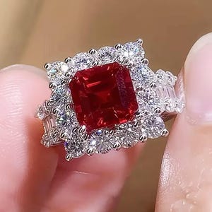 May include: A dazzling ring featuring a large, square-cut, deep red gemstone as the centerpiece. The red stone is surrounded by a halo of smaller, sparkling white gemstones. The ring's band is also adorned with additional white gemstones, creating a luxurious and eye-catching design.