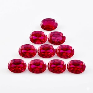 May include: A collection of eleven oval-cut, vibrant red gemstones arranged in a triangular pattern against a white background. The gems exhibit a high degree of clarity and brilliance, reflecting light to create a sparkling effect. These gemstones are suitable for jewelry making.