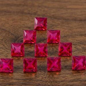 May include: Ten square-cut, vibrant pink gemstones arranged on a wooden surface. Each gem has a faceted surface, reflecting light. The gems are a deep, saturated pink color, and are evenly spaced.