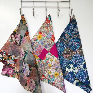 May include: Three colourful patchwork fabric tea towels hanging on a silver rack. The towels are made of different fabrics with floral and geometric patterns. The towels are in shades of blue, pink, green, and brown.