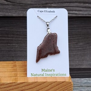 May include: A brown pendant necklace in the shape of the state of Maine, displayed on a white card that reads "Cape Elizabeth." The card is propped up on a wooden stand, with the text "Maine's Natural Inspirations" below. The necklace is on a silver chain.