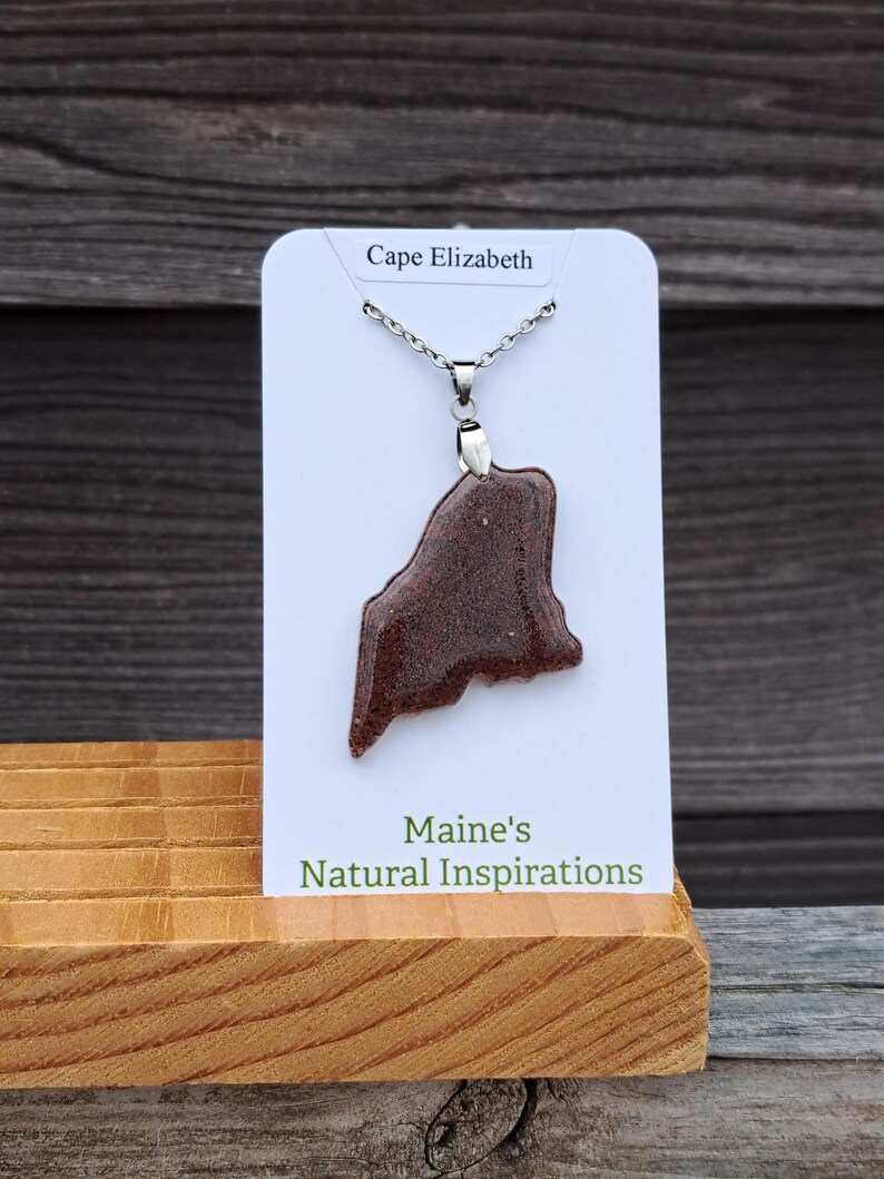 May include: A brown pendant necklace in the shape of the state of Maine, displayed on a white card that reads "Cape Elizabeth." The card is propped up on a wooden stand, with the text "Maine's Natural Inspirations" below. The necklace is on a silver chain.
