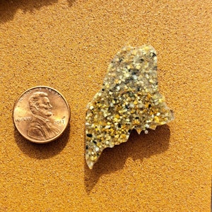 May include: A resin-filled state of Maine outline, shaped like the state, with a sandy, speckled texture. The outline is about the size of a US penny.