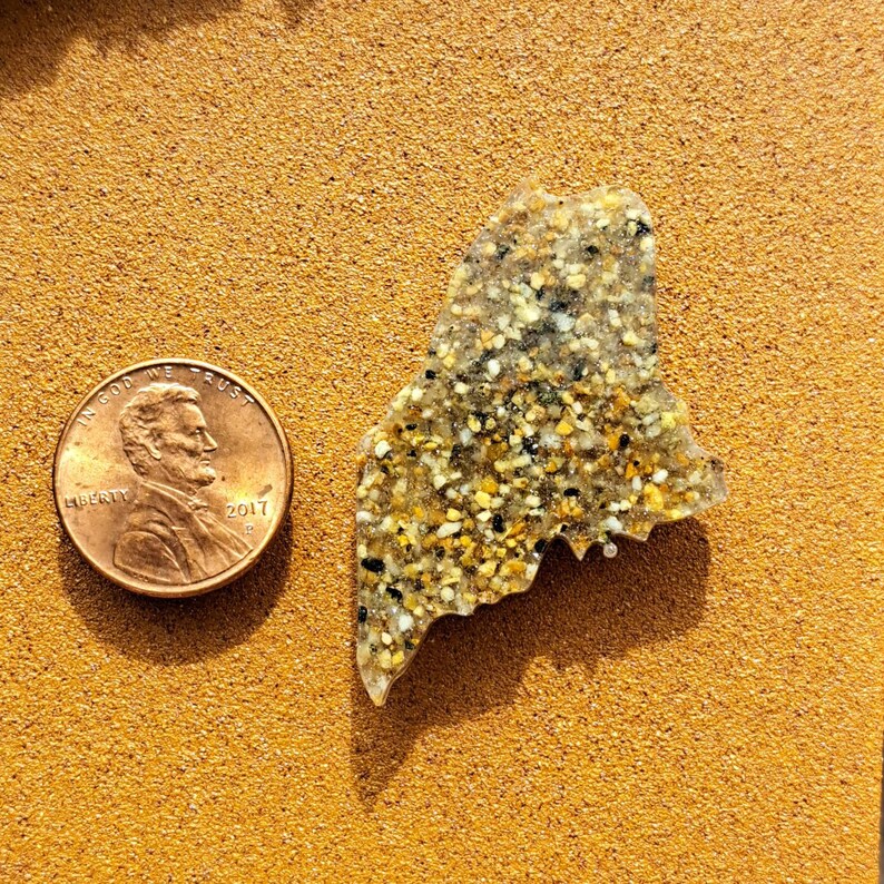 May include: A resin-filled state of Maine outline, shaped like the state, with a sandy, speckled texture. The outline is about the size of a US penny.