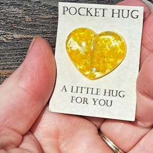 May include: A Pocket Hug card with a yellow heart-shaped charm. The card reads "POCKET HUG" at the top and "A LITTLE HUG FOR YOU" at the bottom. The heart charm is filled with yellow flecks.