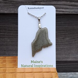 May include: A silver necklace with a pendant shaped like the state of Maine. The pendant is a gray, textured stone. The card behind the necklace reads "Kennebunkport" and "Maine's Natural Inspirations."