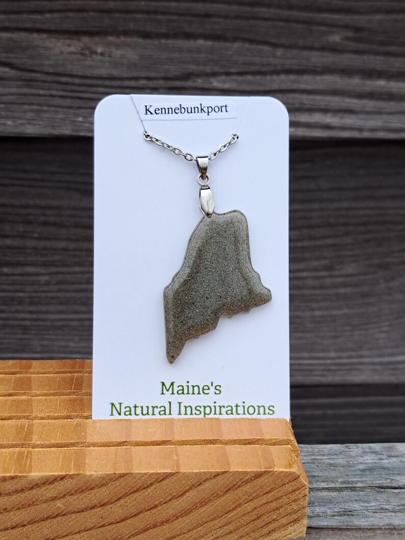 May include: A silver necklace with a pendant shaped like the state of Maine. The pendant is a gray, textured stone. The card behind the necklace reads "Kennebunkport" and "Maine's Natural Inspirations."
