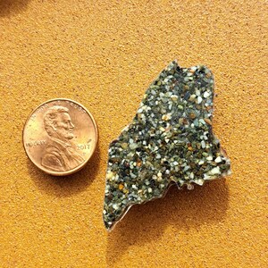 May include: A small, green and brown, resin-like, state-shaped paperweight of Maine. The paperweight is next to a US penny coin with the year 2017.