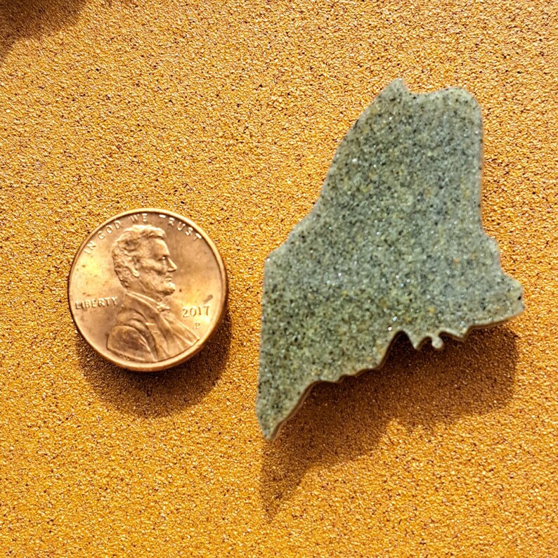 May include: A small, gray, stone-like cutout of the state of Maine. The cutout is about the size of a US penny.